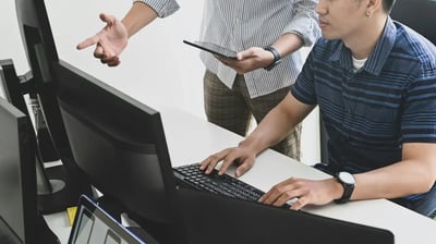 Person working at a computer while a colleague gestures and holds a tablet during an office discussion