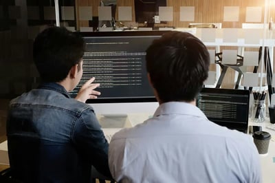 Two developers reviewing code together on desktop and laptop screens in an office workspace