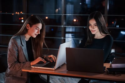 Two women working on laptops together in a modern office at night